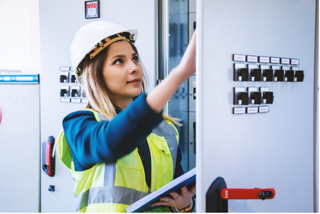 A Hispanic female in safety gear looks over a wiring box