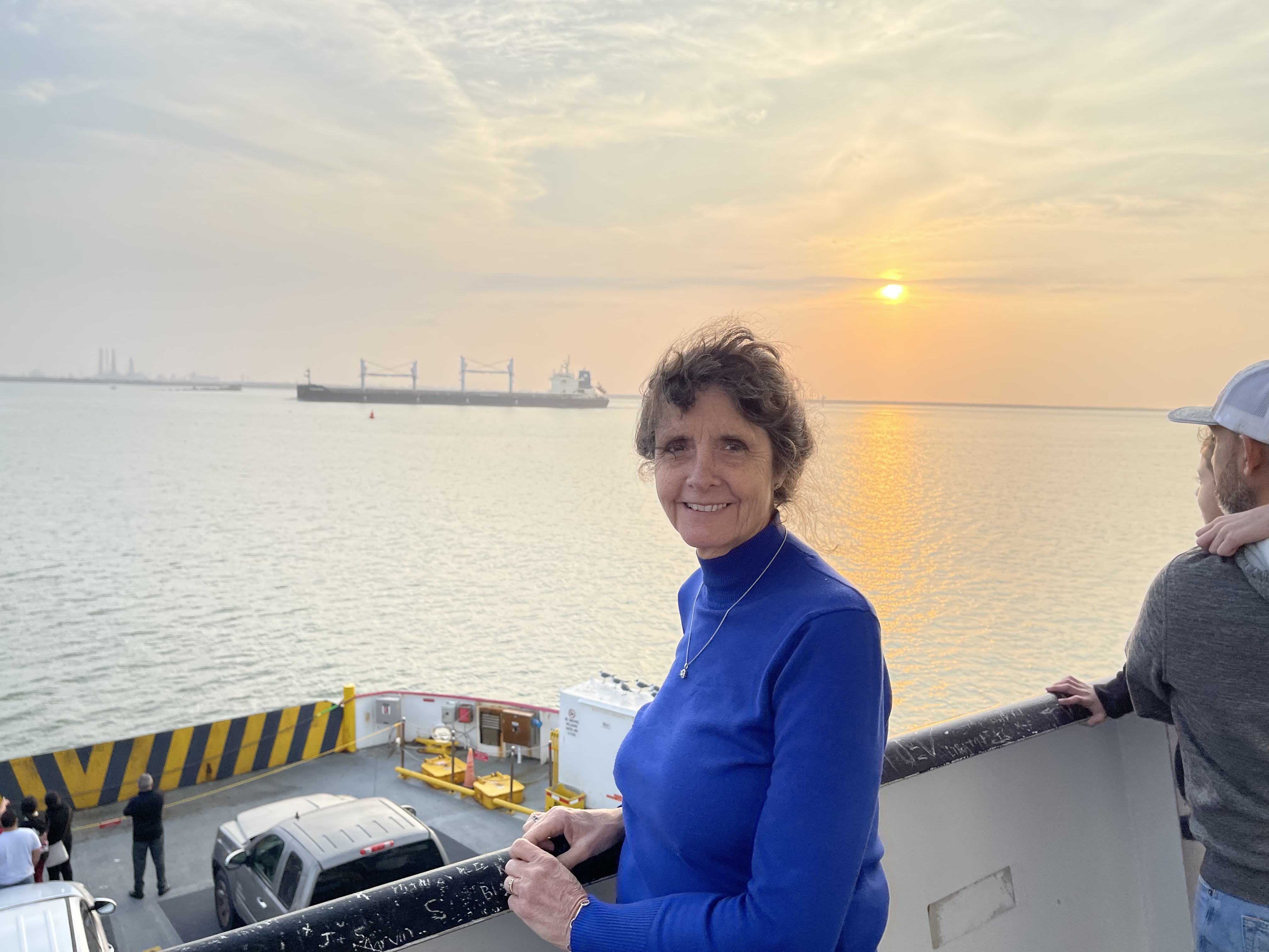 A person in a blue top stands on a ferry deck overlooking calm water at sunset, with a cargo ship in the distance and vehicles parked below.