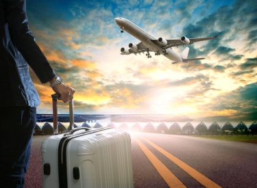 An airport runway with a person standing with luggage and an airplane flying overhead