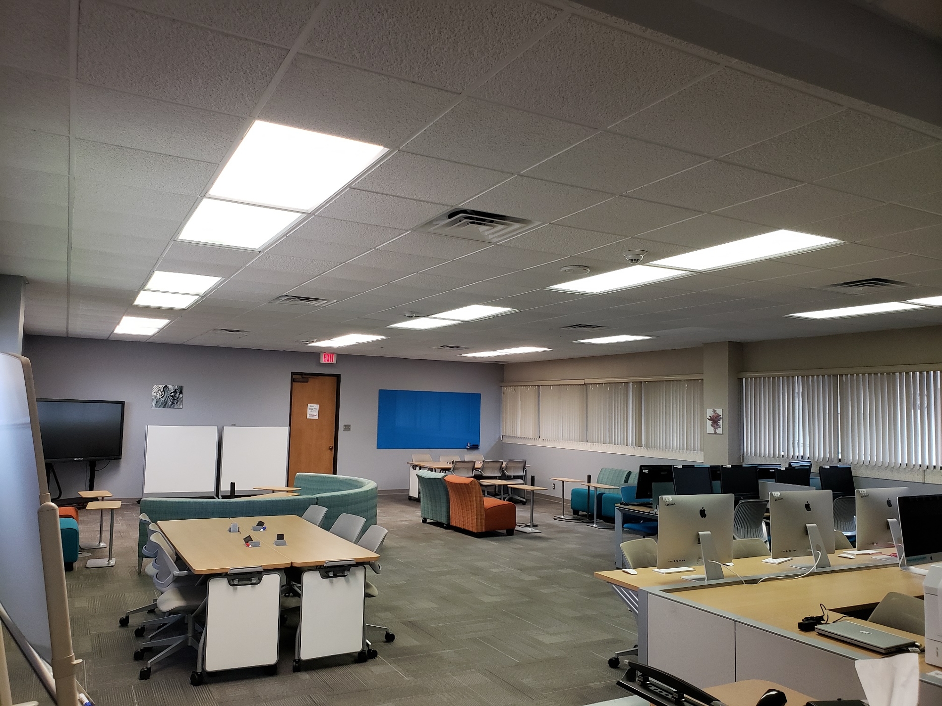 Wide view of main Collaboratory room with various sized tables/chairs, computers, and dry erase boards.