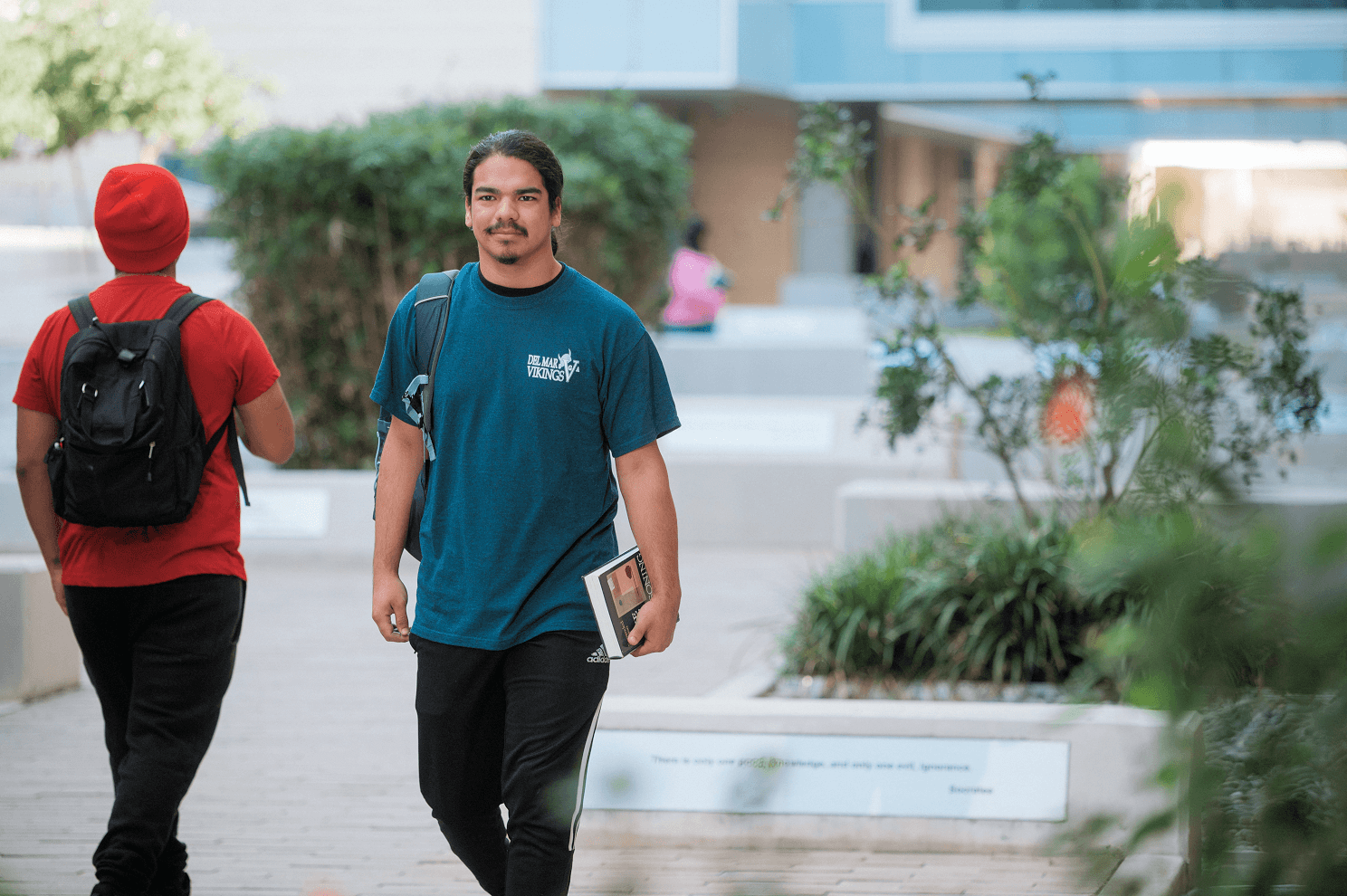 Two students walk through a landscaped campus courtyard; one wears a teal Del Mar Vikings shirt and carries a book, while the other wears red and carries a black backpack.