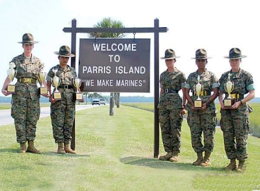 Five people in military uniforms holding trophies stand in front of a "Welcome to Parris Island" sign.